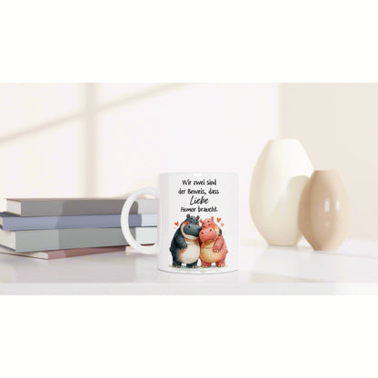 White mug mockup showing two cartoon hippos hugging with hearts and a German caption; sits among books and decorative vases.