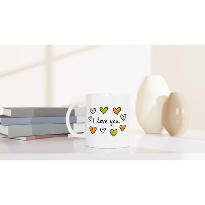 White ceramic mug with I love you and colorful hearts, placed on a desk beside books and decorative beige vases.