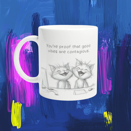 White ceramic mug with a sketch of two laughing cats and the caption 'You're proof that good vibes are contagious' against a blue abstract background.