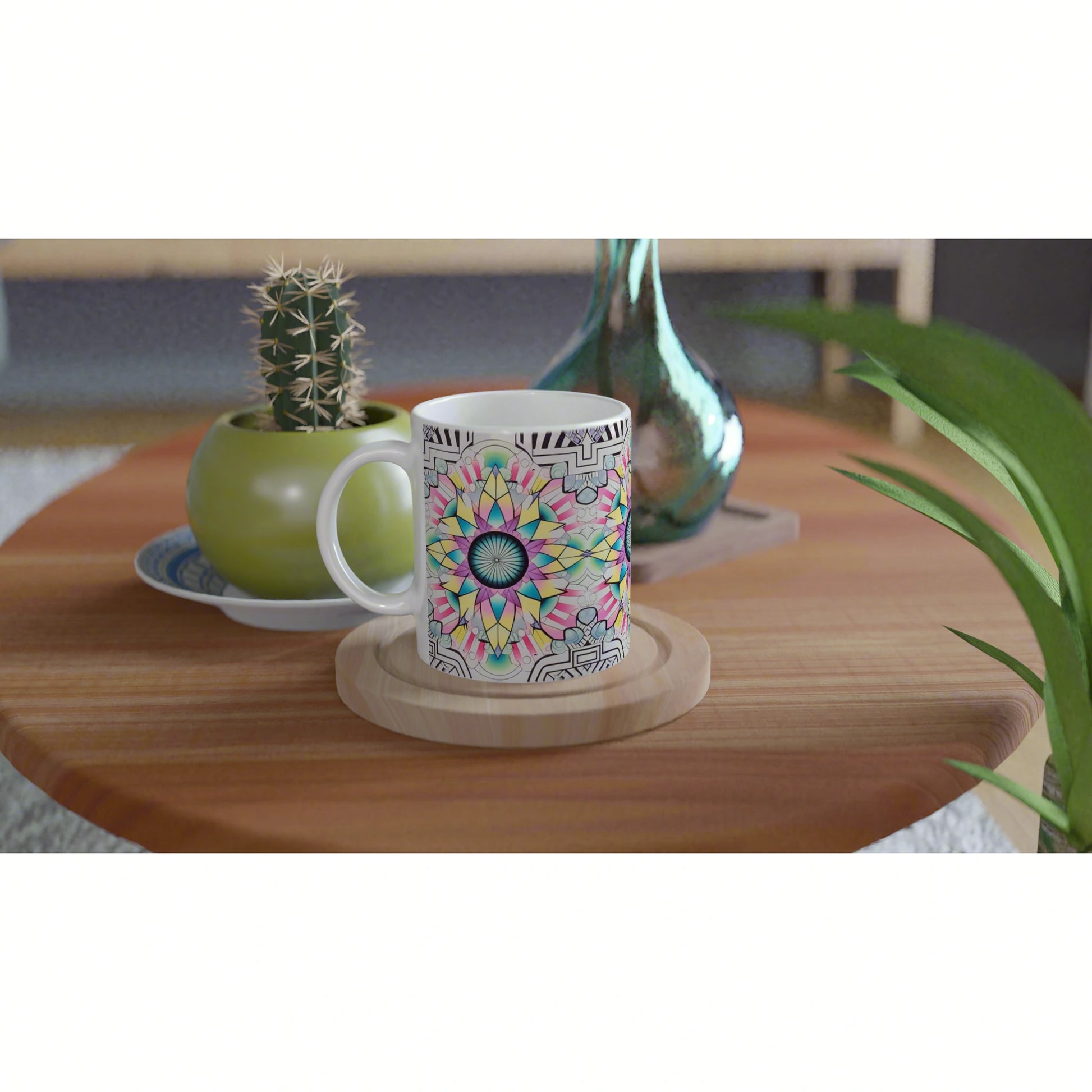 Product mockup: white mug with colorful mandala design on a coaster, on a wooden table with a cactus plant and vase nearby.
