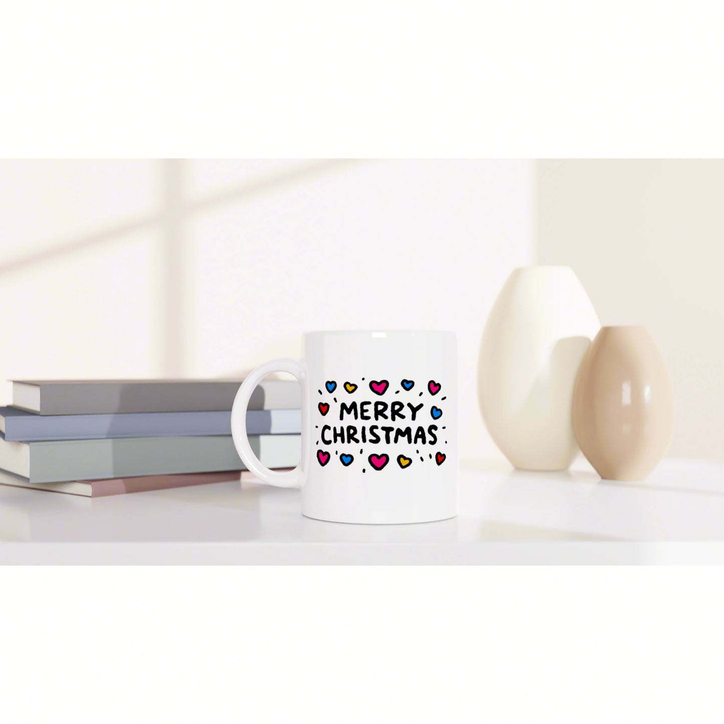 White mug with colorful hearts and the text MERRY CHRISTMAS, placed on a desk beside books and decorative vases.