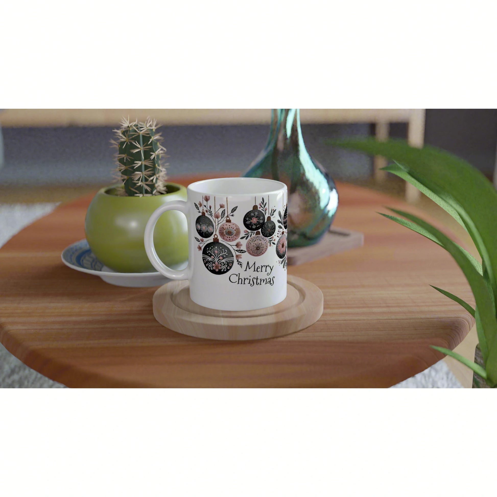 White mug with Merry Christmas and ornament motifs on a wooden table beside a small cactus and teal glass vase.