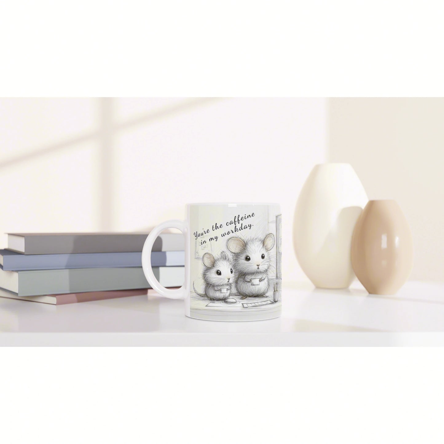 White mug with a cute mouse illustration and the caption 'You're the caffeine in my workday' on a desk beside books and beige vases.