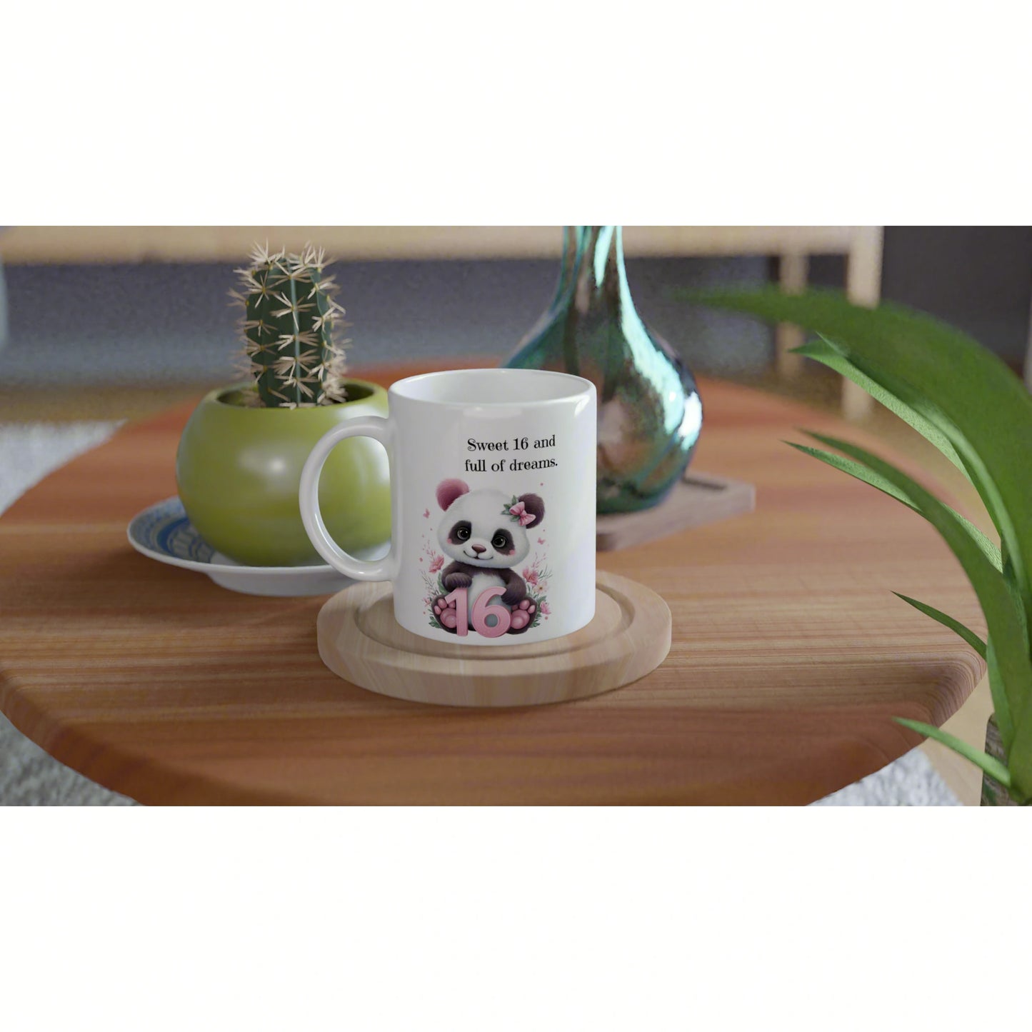 White mug with a panda illustration and the text 'Sweet 16 and full of dreams', set on a wooden coaster with plant decor in the background.
