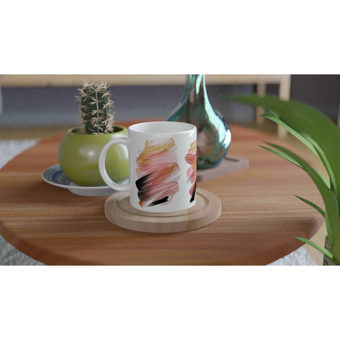 White mug with pink-orange abstract brush strokes on a wooden coaster, placed on a table with a cactus and a green vase.
