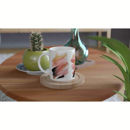 White mug with pink-orange abstract brush strokes on a wooden coaster, placed on a table with a cactus and a green vase.