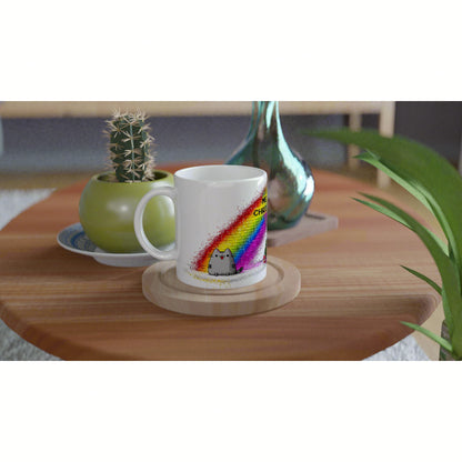 White ceramic mug featuring a rainbow splash and a tiny cartoon cat, displayed as a product mockup on a wooden table.