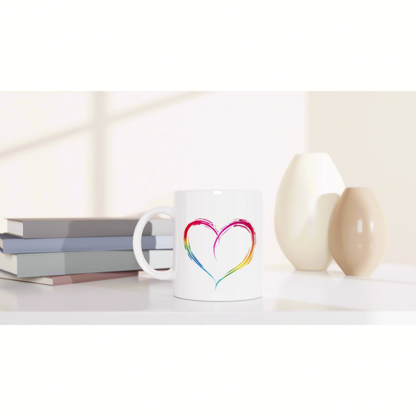 White mug with a rainbow heart design on a white tabletop, beside stacked books and two ceramic vases.