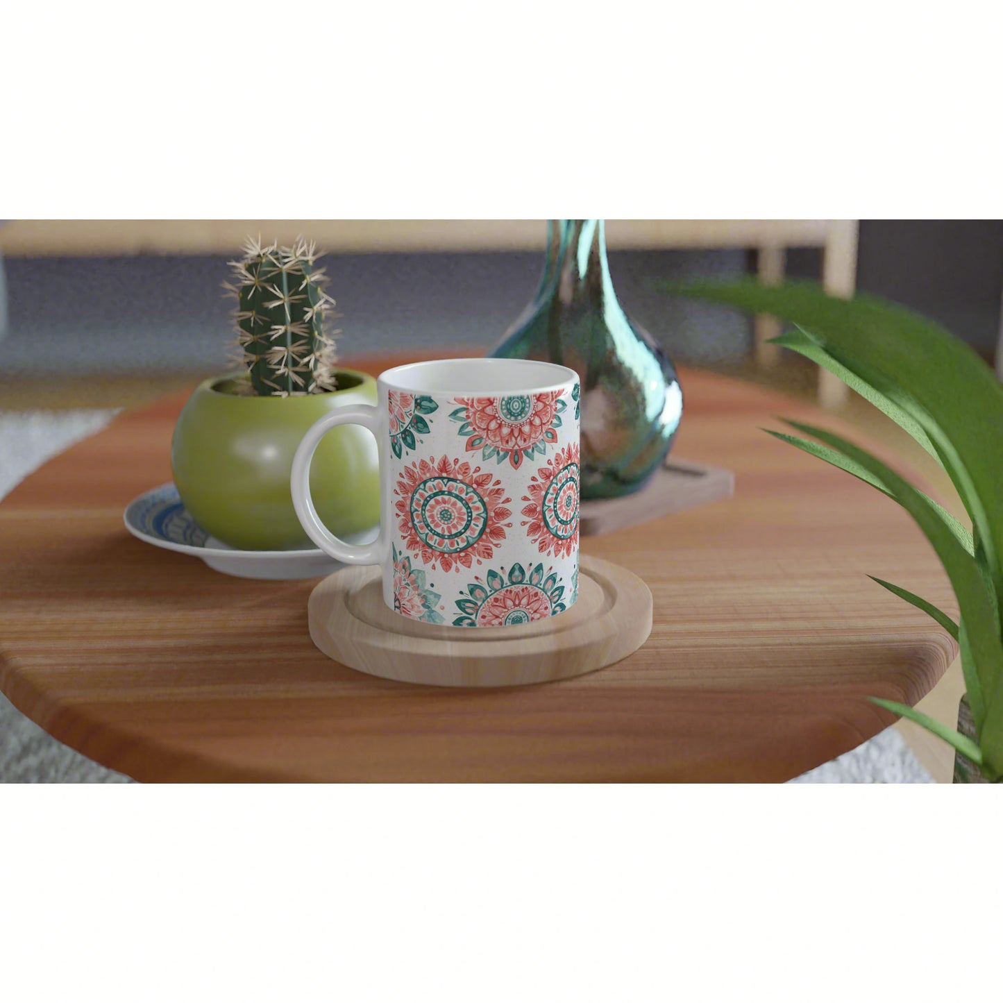 White mug with teal and coral mandala pattern on a wooden coaster, on a wood table with a cactus pot and blue glass vase.