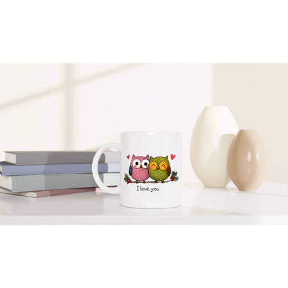 White mug featuring two cartoon owls, hearts, and the I love you phrase, set on a desk with books and vases.