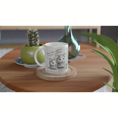White mug with a sketch of two mice and the caption 'You're the caffeine in my weekday' on a wooden coaster, among plants.