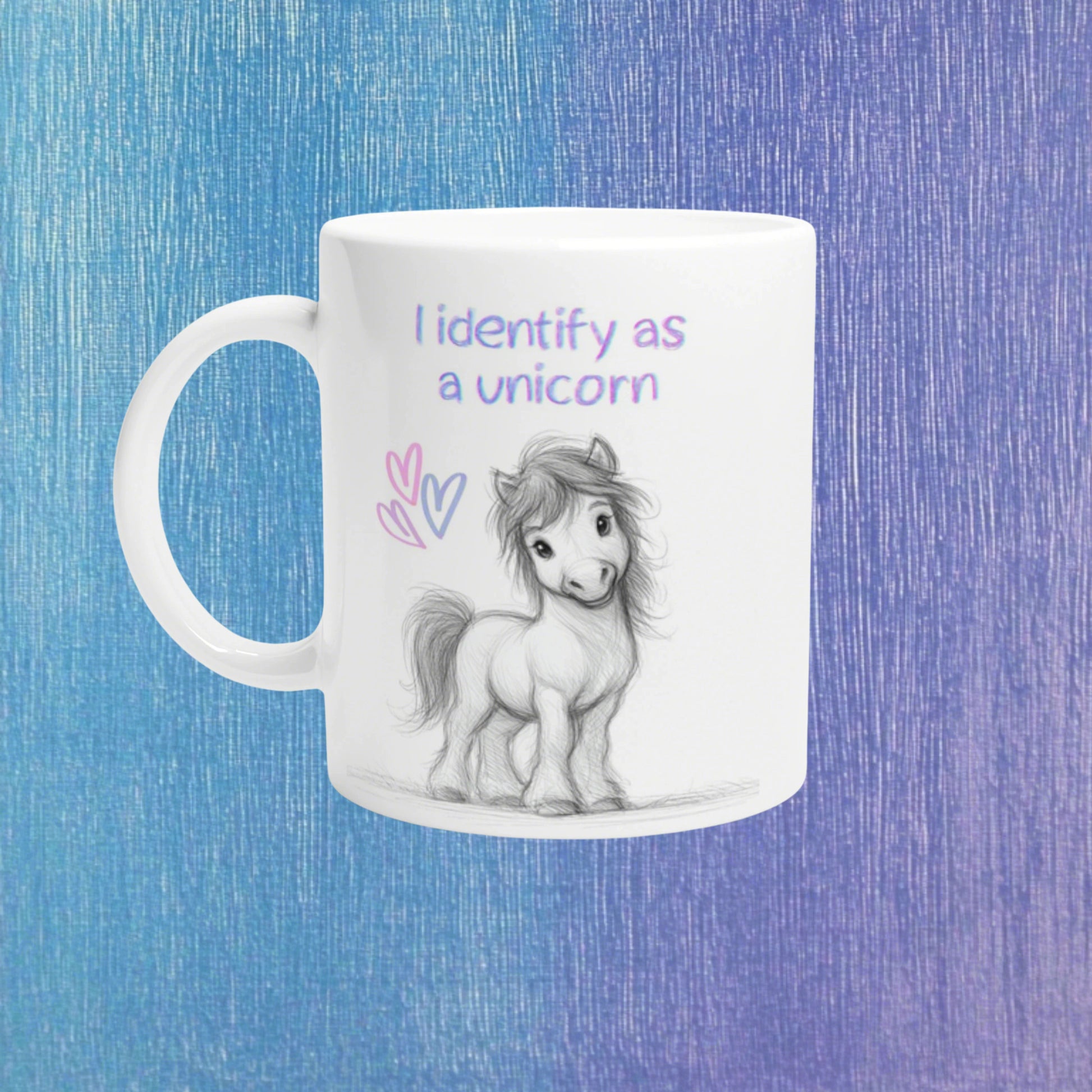 White mug mockup featuring a pencil-drawn unicorn and slogan 'I identify as a unicorn' with pink hearts on a blue gradient background.