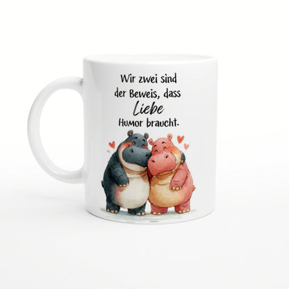 White mug with a watercolor illustration of two hugging hippos, hearts, and a German slogan about love needing humor.