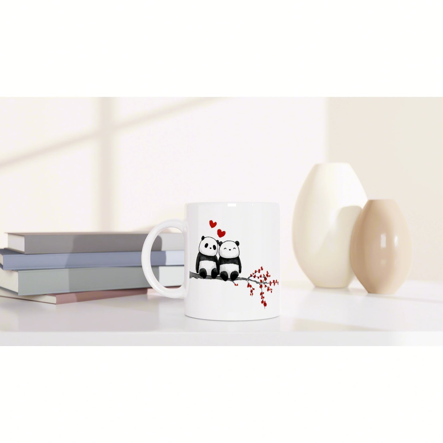 White mug with two pandas perched on a branch with red hearts, set among pastel books and beige vases on a clean desk.