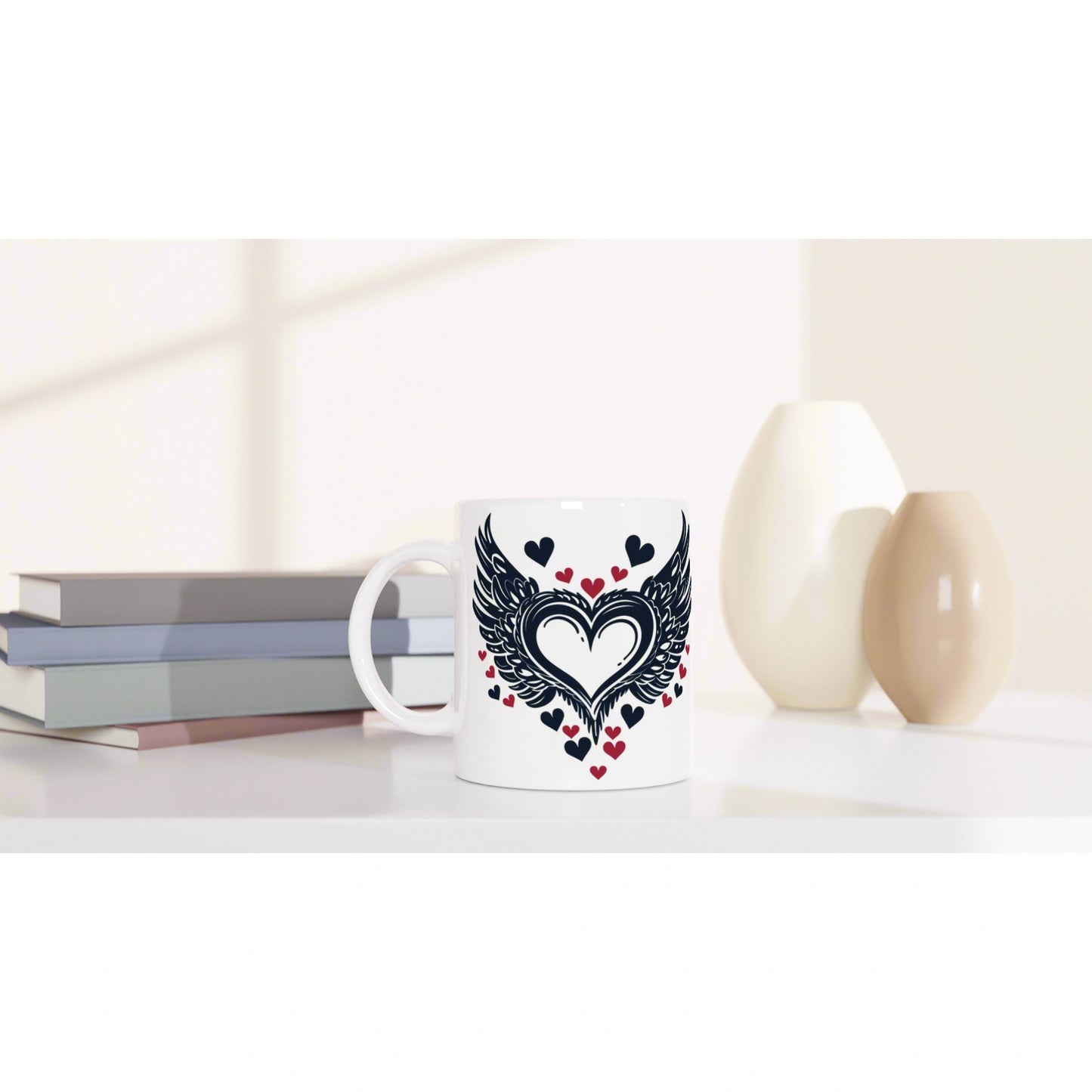 White mug with a winged heart design and red hearts, on a desk with books and beige vases.