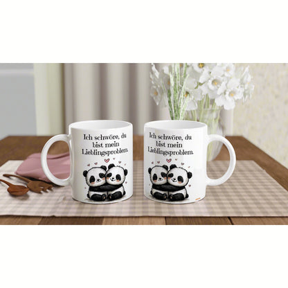 Zwei weiße Tassen mit Panda-Paar und dem Spruch 'Ich schwöre, du bist mein Lieblingsproblem' auf einem Holztisch.